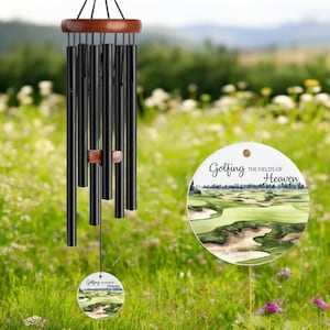 May include: Black wind chimes with a wooden top and accents, hanging in a garden. The chimes have a round, decorative tag with the words "Golfing the Fields of Heaven" and a golf course image.