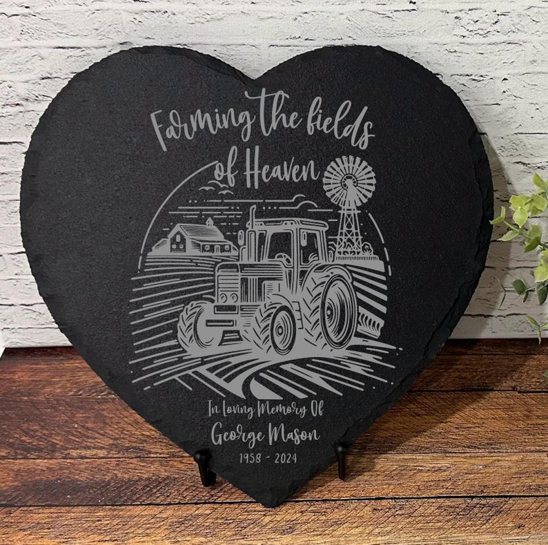 May include: Black heart-shaped slate with a gray engraved image of a tractor in a field with a windmill and the text "Farming the fields of Heaven". The text "In loving memory of George Mason 1958 - 2024" is also engraved on the slate.