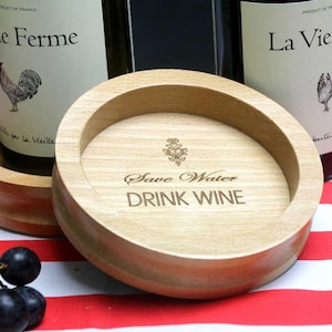 May include: A round wooden coaster with a raised edge. The coaster is light brown and features a laser-engraved design that reads "Save Water Drink Wine".