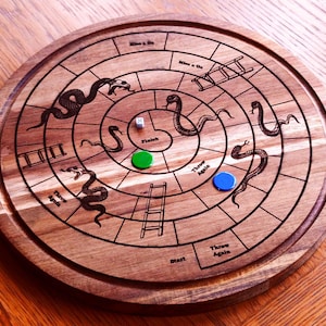 May include: A wooden Snakes and Ladders board game. The circular board has a spiral design with snakes, ladders, and game spaces. A green and blue game piece and a die are on the board. The board is on a wooden surface.