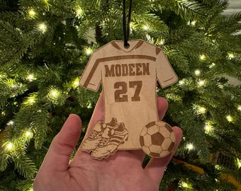 May include: A wooden soccer-themed Christmas ornament. The ornament is shaped like a jersey with the name "MODEEN" and the number "27". It also features soccer cleats and a soccer ball. The ornament is hanging in front of a Christmas tree.