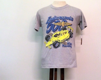 May include: A gray t-shirt featuring a yellow race car and the text "Indianapolis 500". The shirt also has the text "PENZOIL 731 Running" and a graphic of a winged emblem. The shirt is displayed on a wooden stand.