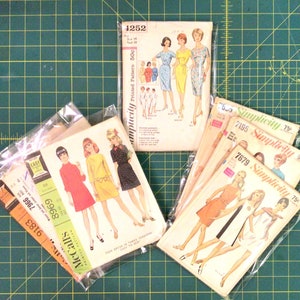 May include: A collection of vintage sewing patterns, including Simplicity and McCall's brands, featuring illustrations of dresses. The patterns are in plastic sleeves and are on a green grid surface. Some patterns have prices like 50¢ and 75¢.