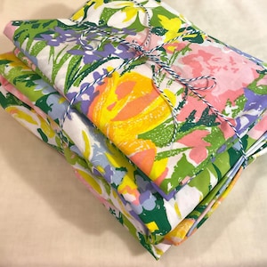 May include: A stack of folded fabric featuring a vibrant floral pattern with large pink, yellow, green, and purple blossoms on a white background. The fabric is tied together with a thin blue and white baker's twine string wrapped around the center.