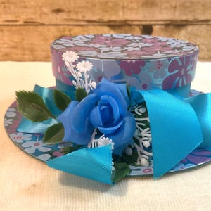 Puede incluir: Una pequeña caja para sombreros redonda y de tapa plana con un estampado floral en tonos morados y azules. La base está decorada con una cinta de satén turquesa brillante, una rosa azul y pequeñas ramitas de flores blancas con hojas verdes.