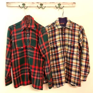 Puede incluir: Dos camisas de cuadros de manga larga colgadas en la pared. Una es verde y roja, la otra una mezcla de amarillo, azul y blanco. Ambas tienen botones y cuellos. Estilo vintage, con un dise&ntilde;o cl&aacute;sico y atemporal.