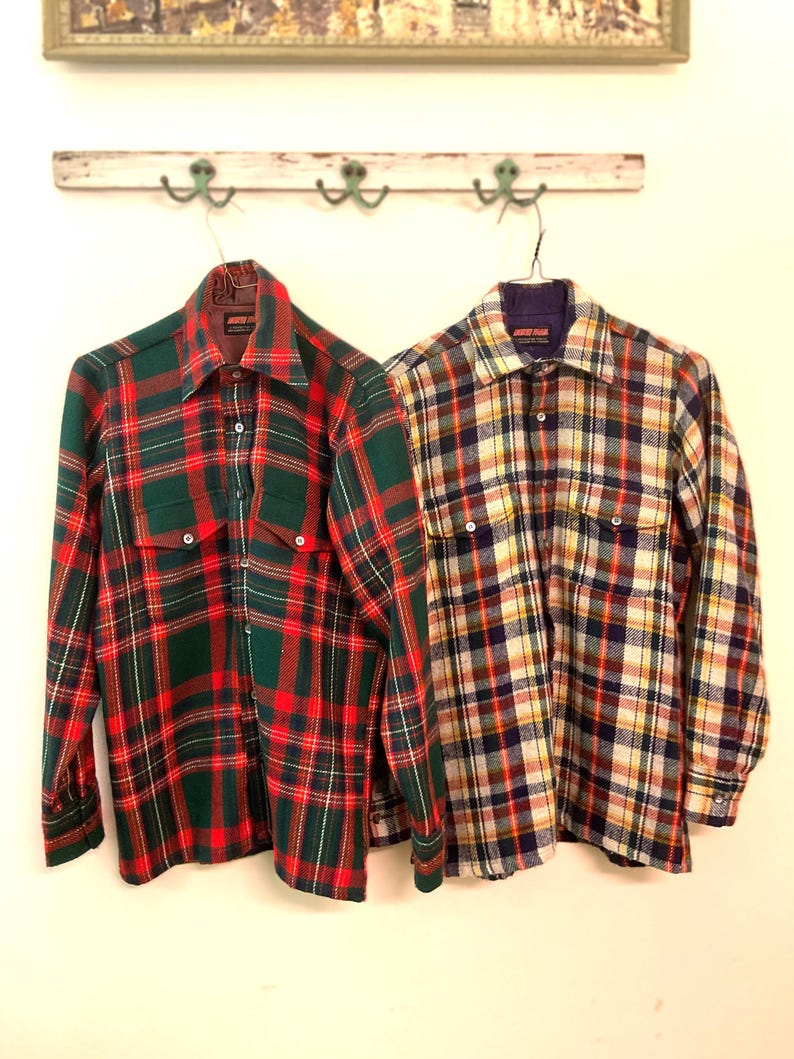 Puede incluir: Dos camisas de cuadros de manga larga colgadas en la pared. Una es verde y roja, la otra una mezcla de amarillo, azul y blanco. Ambas tienen botones y cuellos. Estilo vintage, con un dise&ntilde;o cl&aacute;sico y atemporal.