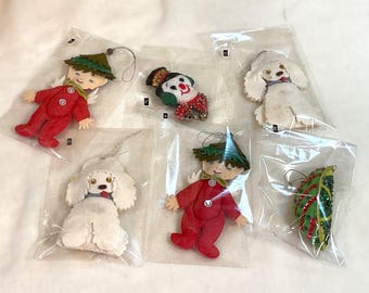 Könnte beinhalten: Eine Sammlung von Vintage-Weihnachtsschmuck in durchsichtigen Kunststoffverpackungen. Die Ornamente umfassen einen rot gekleideten Elf, einen weißen Pudel, einen Schneemann und ein grünes Blatt, alle mit dekorativen Details und Aufhängeschlaufen.