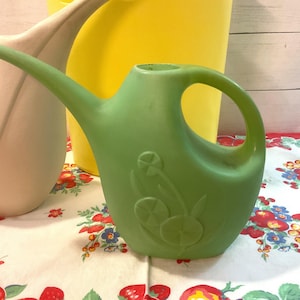 May include: A green plastic watering can with a curved handle and a long spout. The watering can has a floral design on the side. A pink and yellow watering can are in the background. The watering cans are on a floral patterned tablecloth.