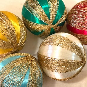 Könnte beinhalten: Eine Sammlung dekorativer Kugeln in verschiedenen Farben, darunter Gelb, Grün, Blau, Weiß und Rot. Jede Kugel ist mit goldfarbenen, fadenartigen Verzierungen versehen, die ein festliches und elegantes Aussehen erzeugen. Geeignet für Weihnachtsdekorationen.