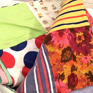 Op de afbeelding: Een kleurrijke patchworkquilt met verschillende stofpatronen. De quilt heeft een groene vierkant, een witte stof met rode en blauwe stippen en een bloemenprint. Andere patronen zijn strepen en nautische motieven.