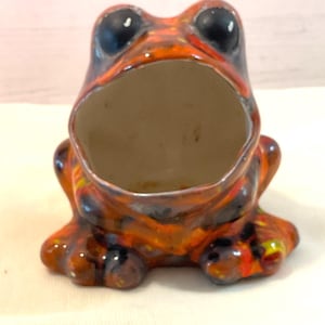 May include: A ceramic frog figurine with a wide open mouth, painted in shades of orange, black, and yellow. The frog has large black eyes and a glossy finish. The figurine is a decorative item.
