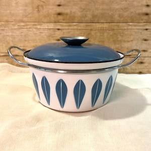 Könnte beinhalten: Ein weißer Emailletopf mit passendem blauem Deckel und zwei seitlichen Griffen. Die Außenseite des Topfes ist mit einem sich wiederholenden Muster aus vertikalen, blauen blattförmigen Motiven verziert. Der Rand ist mit einer schwarzen Kante.