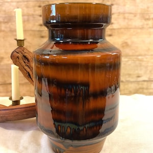 May include: A vintage ceramic vase with a glossy, layered glaze in shades of brown, orange, and black. The vase has a cylindrical body with a flared base and a slightly wider top. The background includes a wooden surface and a candle holder.