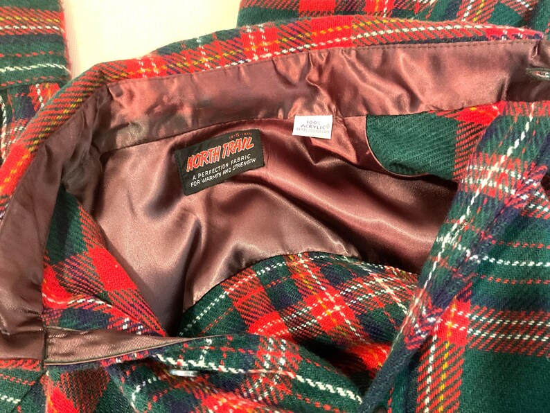 Puede incluir: Primer plano de una chaqueta a cuadros con patrones rojos, verdes y blancos. La chaqueta tiene un forro de sat&eacute;n marr&oacute;n y una etiqueta que dice "NORTH TRAIL A PERFECTION FABRIC FOR WARMTH AND STRENGTHEN."