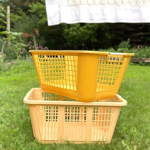 May include: Two rectangular plastic storage baskets stacked vertically. The top basket is a vibrant mustard yellow, while the bottom basket is a lighter, pale peach color. Both baskets feature a grid-like open weave design on all sides.