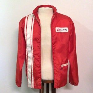 May include: A red jacket with a white stripe down the left side and a small "Simplicity" logo on the right chest. The jacket has a zipper closure and a small pocket. The jacket is displayed on a mannequin.