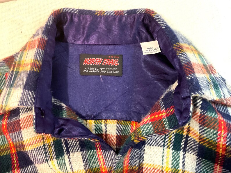 Puede incluir: Primer plano de una camisa de cuadros vintage con forro y cuello de sat&eacute;n morado. La camisa presenta un colorido estampado de cuadros en rojo, verde, amarillo y blanco. La etiqueta dice "NORTH TRAIL" y "100% Acrylic".