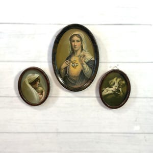 May include: Three oval framed religious prints. The largest depicts a woman with a glowing heart, the second shows a woman holding a baby, and the third shows a cherub with a bow and arrow. The frames are dark brown.