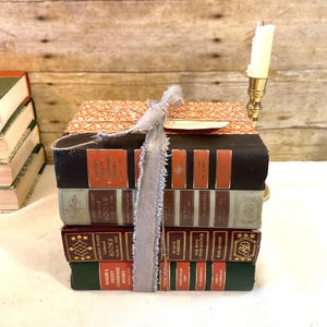 Könnte beinhalten: Ein Stapel alter Bücher, die mit einem grauen Band zusammengebunden sind. Die Bücher haben verschiedene Farben, darunter Schwarz, Grün und Burgunder, mit orangefarbenen und goldenen Buchstaben. Eine Kerze in einem goldenen Kerzenhalter ist zu sehen. Die Bücher sind Reader's Digest Condensed Books.