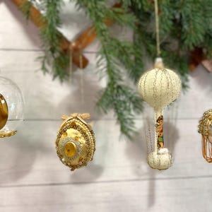 May include: Four vintage Christmas ornaments hanging against a white wood background. The ornaments include a clear glass ball with a gold angel, a gold and yellow egg-shaped ornament, a cream-colored ornament with a painted design, and a gold and cream ornament with tassels.
