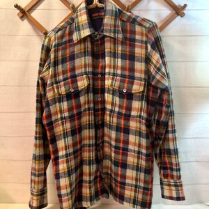 Puede incluir: Una camisa de cuadros de manga larga en una mezcla de colores, incluyendo rojo, azul, amarillo y crema. La camisa tiene dos bolsillos en el pecho y est&aacute; abotonada. Cuelga de un perchero de madera sobre un fondo blanco.
