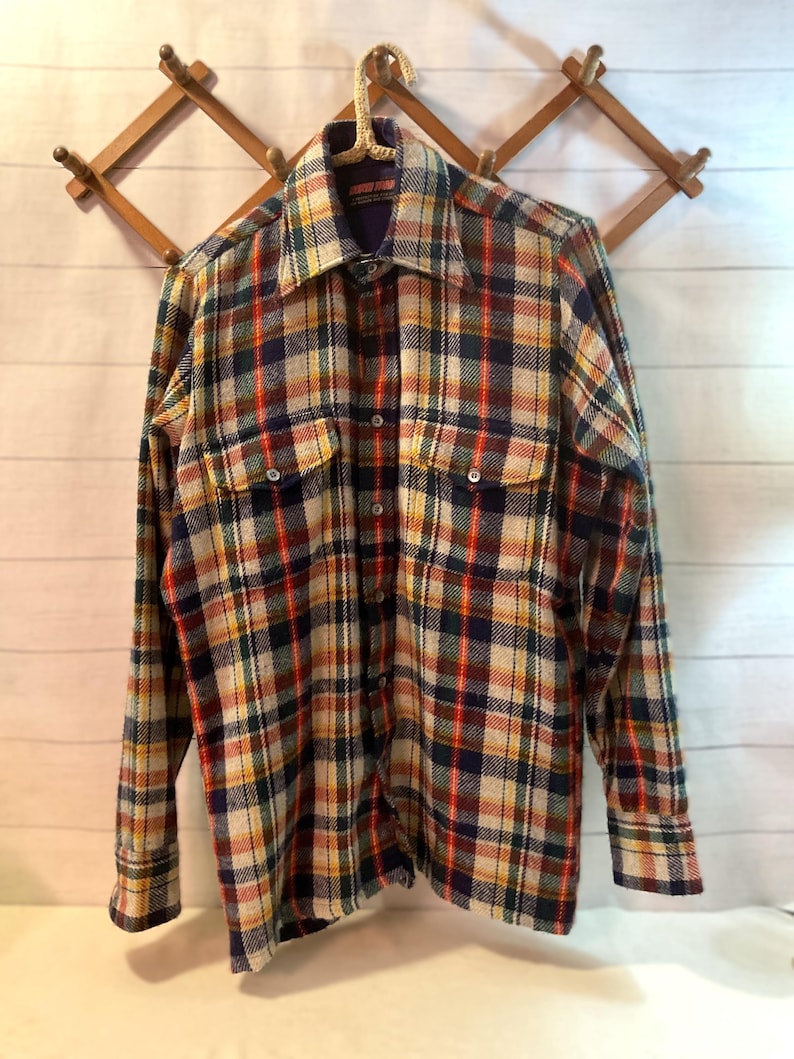 Puede incluir: Una camisa de cuadros de manga larga en una mezcla de colores, incluyendo rojo, azul, amarillo y crema. La camisa tiene dos bolsillos en el pecho y est&aacute; abotonada. Cuelga de un perchero de madera sobre un fondo blanco.