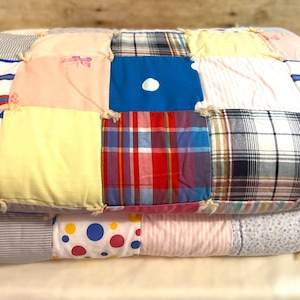 May include: A folded patchwork quilt featuring a variety of fabric squares in diverse patterns and colors, including blue with white polka dots, red and blue plaid, solid yellow, light pink, and striped designs, all joined with visible knotted ties.