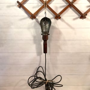 Puede incluir: Una lámpara de estilo vintage con una pantalla de jaula de metal oscuro y un mango de madera. La lámpara está suspendida de un perchero de madera. El cable eléctrico está enrollado y atado con hilo. El fondo es una pared de madera blanca.