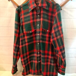 Puede incluir: Una camisa de cuadros de manga larga con un patr&oacute;n cl&aacute;sico rojo, verde y blanco. La camisa tiene dos bolsillos en el pecho, una botonadura y un cuello ancho. La camisa est&aacute; colgada en un perchero de madera.