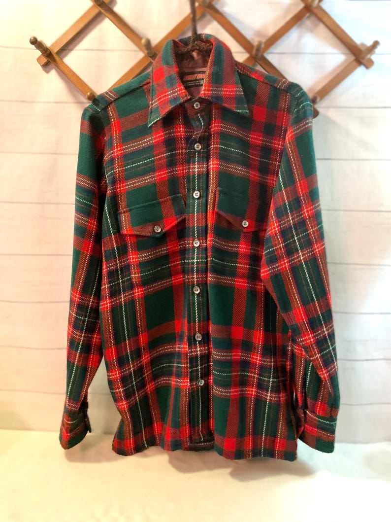 Puede incluir: Una camisa de cuadros de manga larga con un patr&oacute;n cl&aacute;sico rojo, verde y blanco. La camisa tiene dos bolsillos en el pecho, una botonadura y un cuello ancho. La camisa est&aacute; colgada en un perchero de madera.