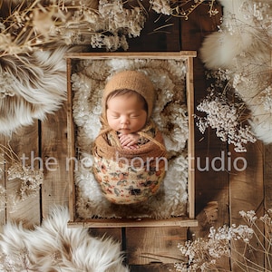 May include: A newborn baby wrapped in a floral patterned blanket and wearing a brown knitted cap, nestled in a wooden box. The box sits on a wooden surface, surrounded by dried flowers and fluffy white fabric. The text "the newborn studio" is visible.