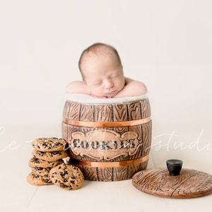 May include: A newborn baby rests inside a decorative wooden cookie jar with the word "COOKIES" on the front. A stack of chocolate chip cookies sits next to the jar, and the lid is off to the side.