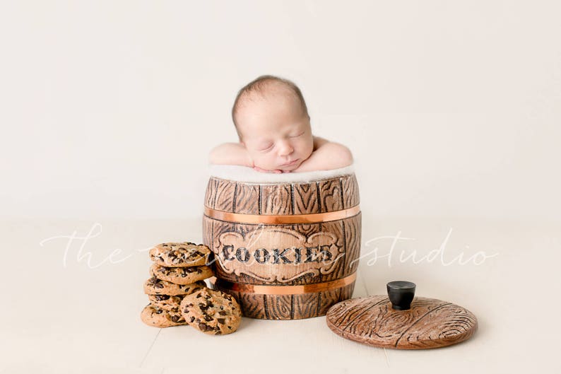May include: A newborn baby rests inside a decorative wooden cookie jar with the word "COOKIES" on the front. A stack of chocolate chip cookies sits next to the jar, and the lid is off to the side.