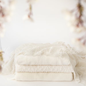 Può includere: Una pila di tre coperte piegate color crema, una con un motivo strutturato e un tessuto di pizzo drappeggiato. Sfondo sfocato con rami floreali appesi. The Newborn Studio è scritto in corsivo sul tessuto.