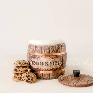 May include: A wooden cookie jar with a copper band and the word "COOKIES" in the center. The jar is open, with a white fluffy material inside. A stack of chocolate chip cookies sits next to the jar, and the lid is to the side.