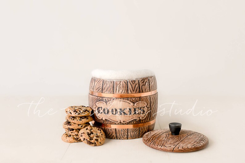 May include: A wooden cookie jar with a copper band and the word "COOKIES" in the center. The jar is open, with a white fluffy material inside. A stack of chocolate chip cookies sits next to the jar, and the lid is to the side.