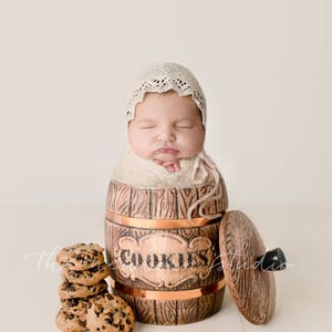 May include: A newborn baby swaddled in a cream-colored blanket and wearing a lace bonnet, nestled inside a wooden cookie jar. A stack of chocolate chip cookies sits beside the jar, with the word "COOKIES" on the jar.
