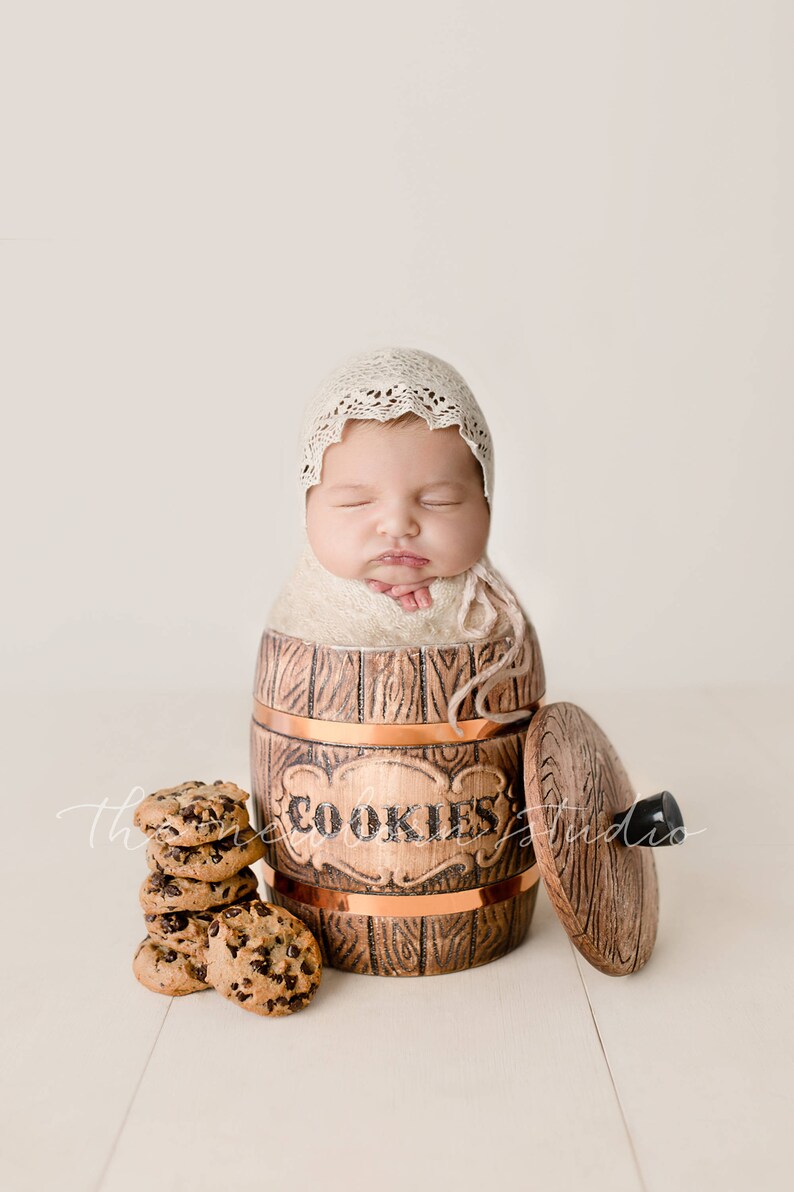 May include: A newborn baby swaddled in a cream-colored blanket and wearing a lace bonnet, nestled inside a wooden cookie jar. A stack of chocolate chip cookies sits beside the jar, with the word "COOKIES" on the jar.