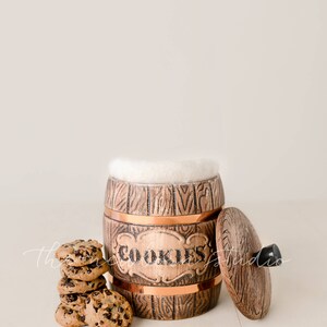May include: A wooden cookie jar with copper accents and the word "COOKIES" on the front. The lid is off, revealing a white interior. A stack of chocolate chip cookies sits beside the jar.