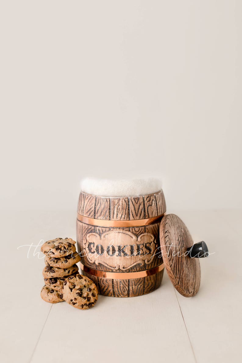 May include: A wooden cookie jar with copper accents and the word "COOKIES" on the front. The lid is off, revealing a white interior. A stack of chocolate chip cookies sits beside the jar.
