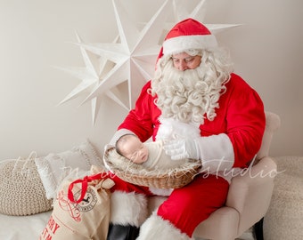 May include: Santa Claus in a red suit and hat, holding a newborn baby in a wicker basket. A Christmas sack with the words "Merry Christmas" is on the left. The background includes white star decorations and a neutral-toned chair.