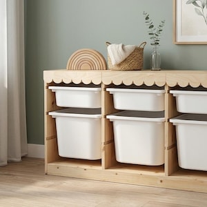 May include: A light-coloured wooden storage unit featuring a scalloped top edge and six white plastic bins arranged in two rows. A woven basket containing a white cloth and a wooden rainbow-shaped toy are placed on the top surface of the unit.