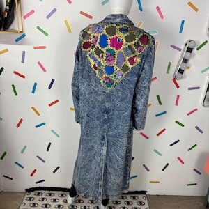 Op de afbeelding: Een lange, zuur gewassen spijkerjas met een kleurrijk, geborduurd ontwerp op de rug. Het ontwerp heeft bloemen en geometrische patronen in roze, blauwe, paarse en gouden tinten. De jas wordt op een mannequin getoond.