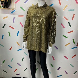 May include: A gold sequin long-sleeved shirt with a collared neckline and button-down front. The shirt is paired with black leggings. The mannequin is standing on a rug with an eye pattern.