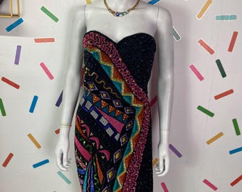 May include: A strapless black cocktail dress with colorful beaded embellishments. The dress features a wrap-style design with geometric patterns in pink, orange, teal, and gold. A gold necklace with colorful stones adorns the mannequin.