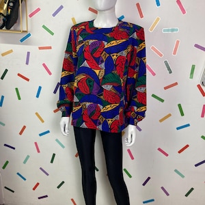 Op de afbeelding: Een lang mouw shirt met een kleurrijke abstracte print in rood, blauw, geel, groen en paars. Het shirt heeft een ronde hals en een losse pasvorm.