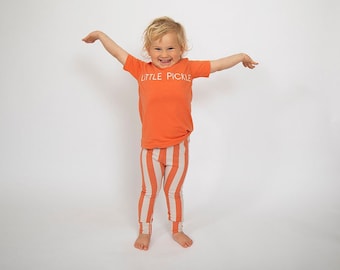 May include: A child wearing an orange t-shirt with the text "LITTLE PICKLE" in white, paired with striped leggings in orange and cream. The child is smiling with arms outstretched, against a white background.
