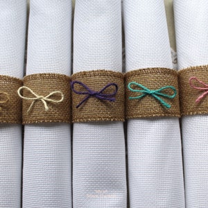 May include: Five white napkins with burlap napkin rings tied with different colored ribbons. The ribbons are brown, cream, purple, teal, and pink.