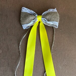 Puede incluir: Un lazo de arpillera con ribete de encaje blanco y cintas de raso amarillo. El lazo está atado con cuerda.